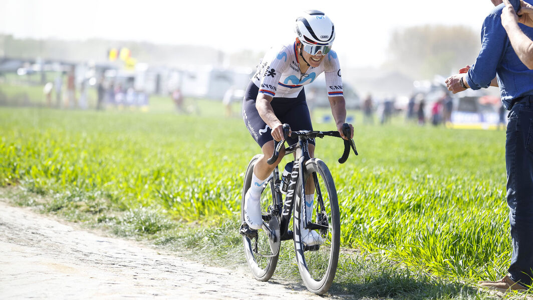 Professional cyclist riding a Canyon road bike on a cobbled race route, wearing team kit and helmet, with spectators in the background.